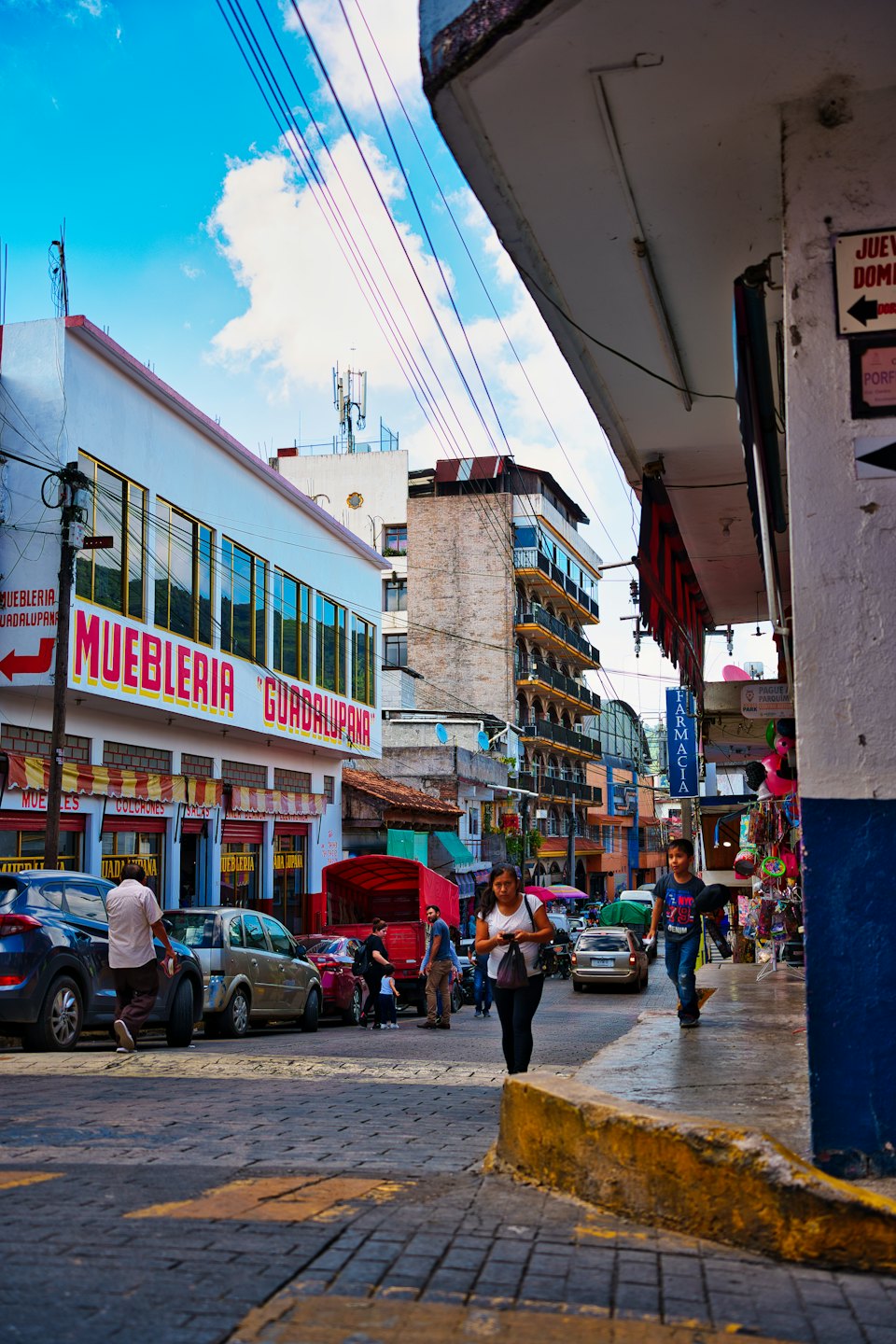Pedestrians walk along a busy cobblestone street past urban storefronts.